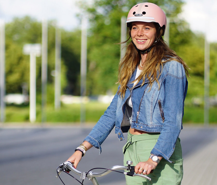 Frau mit braunen Haaren lächelt und steht mit einem Fahrrad und trägt einen rosa Melon E-Series Fahrradhelm »Lotus«.