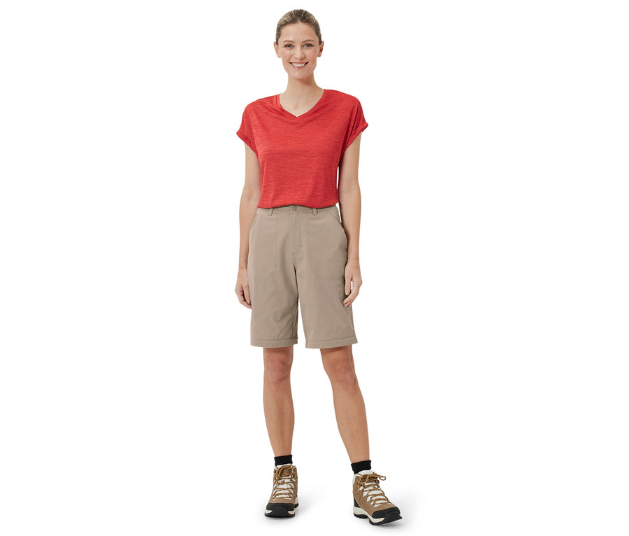 Eine Frau steht und trägt eine beige 2-in-1-Funktionshose, ein rotes T-Shirt und braune Schuhe.