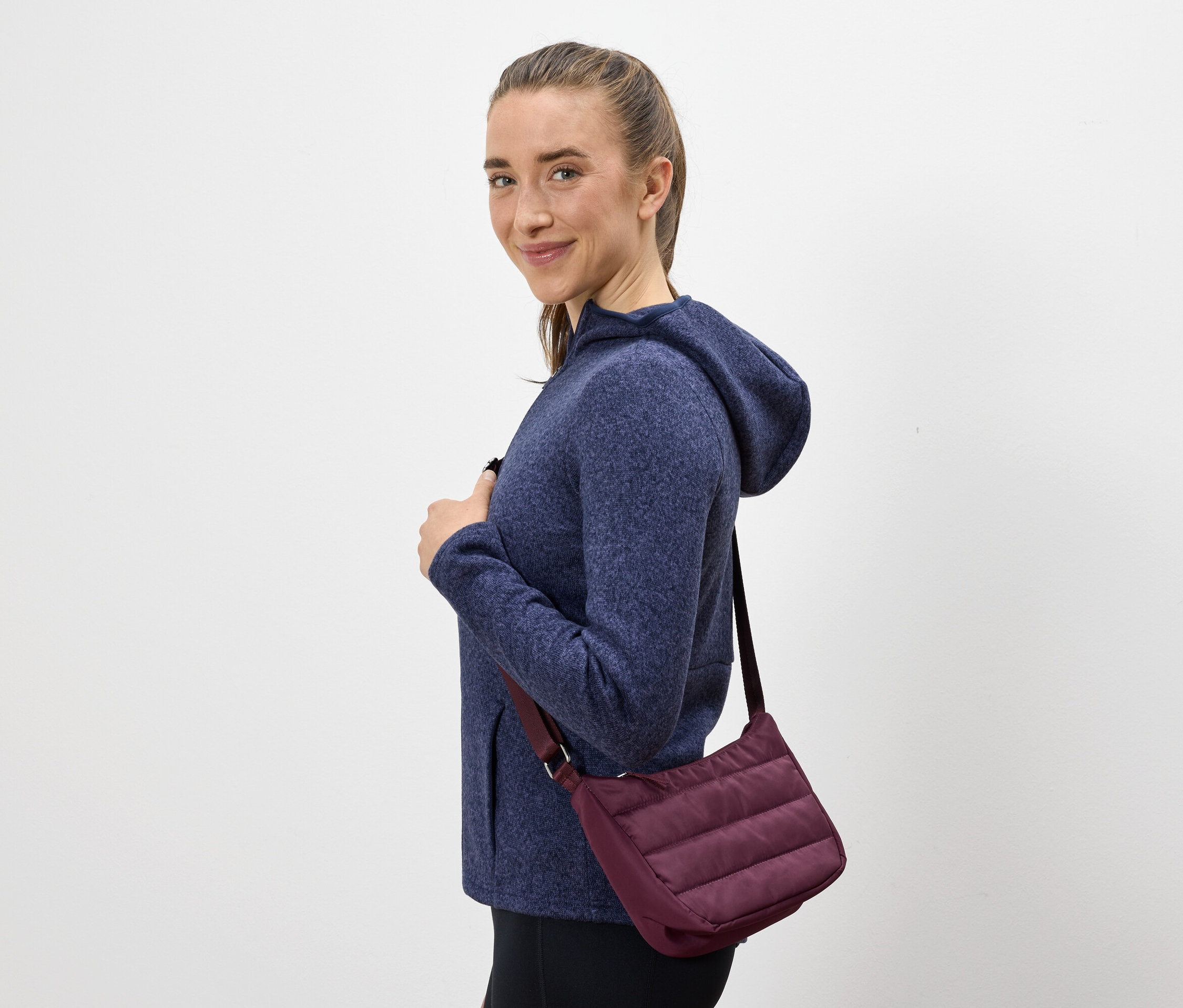 Frau mit braunen Haaren und blauem Kapuzenpullover lächelt über die Schulter. Sie trägt eine violette Handtasche.