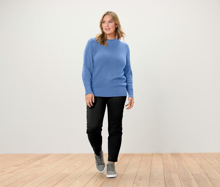 Frau geht auf Holzboden und trägt einen blauen Pullover, schwarze Hose und graue Schuhe.