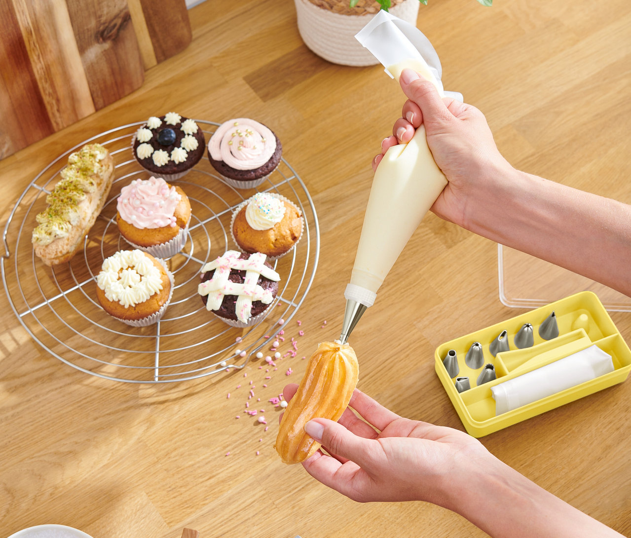 Eine Person dekoriert ein Gebäckstück mit einem Spritzbeutel, Cupcakes und eine gelbe Box mit Tüllen liegen auf einem Holztisch.