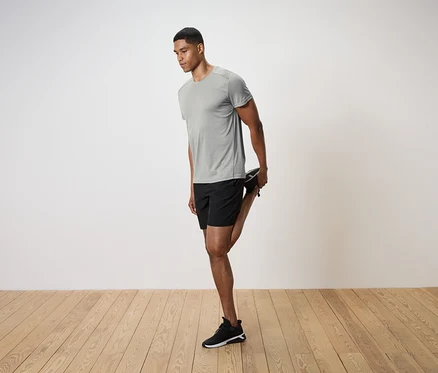 Mann in grauem T-Shirt und schwarzen Shorts dehnt sein Bein auf einem Holzboden.