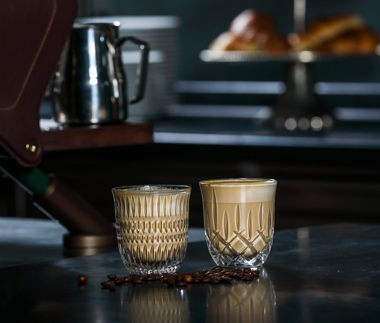 Zwei mit Cappuccino gefüllte Nachtmann »Noblesse« Gläser stehen neben Kaffeebohnen auf einer glänzenden Oberfläche.