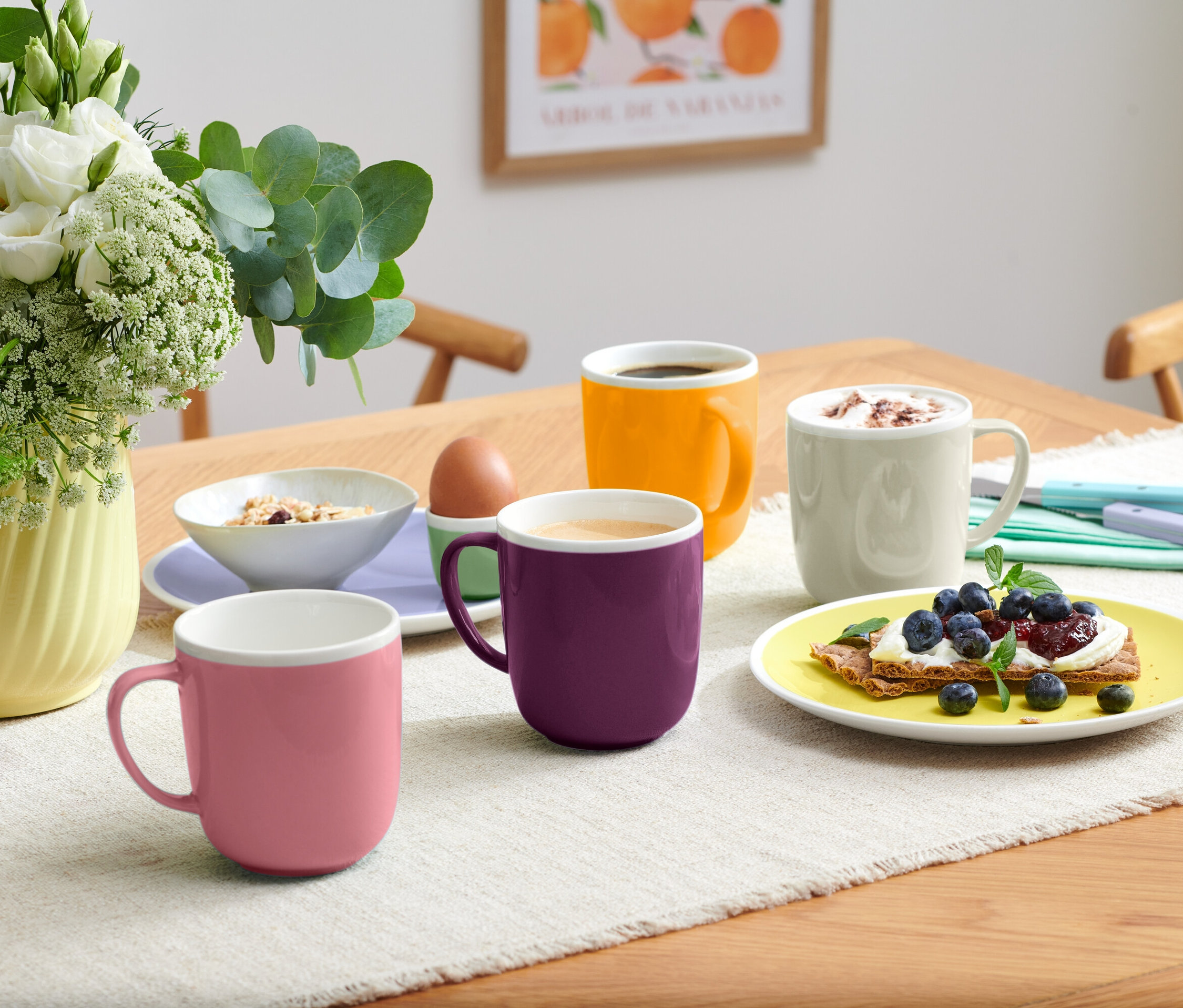 Auf einem Tisch stehen vier verschiedenfarbige Tassen, eine Müslischale, ein Ei im Eierbecher und ein Teller mit einem Blaubeer-Toast.