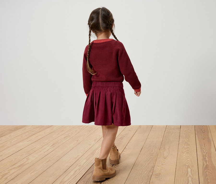 Rückansicht eines Mädchens in bordeauxfarbenem Pullover und Rock, mit braunen Stiefeln, das auf einem Holzboden läuft.