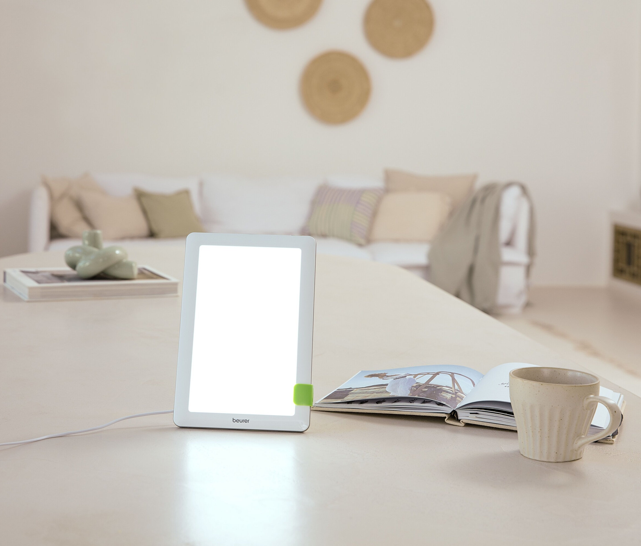 Auf einem Tisch stehen eine Lampe, ein offenes Buch und eine Tasse. Im Hintergrund befindet sich ein Sofa.