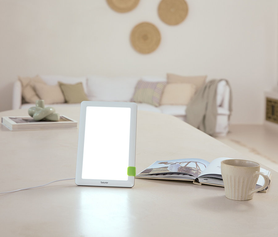 Auf einem Tisch stehen eine Lampe, ein offenes Buch und eine Tasse. Im Hintergrund befindet sich ein Sofa.