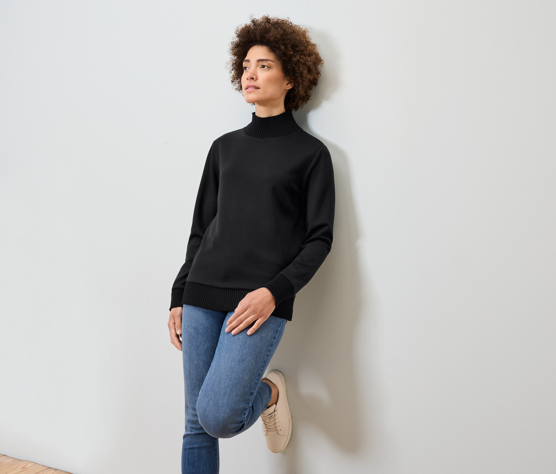 Eine Frau mit Afro-Frisur lehnt an einer weißen Wand. Sie trägt einen schwarzen Rollkragenpullover, blaue Jeans und beige Turnschuhe.