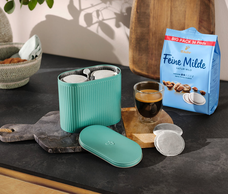 Eine türkisfarbene Pad Box steht neben einer Kaffeepackung und einer Tasse Espresso.