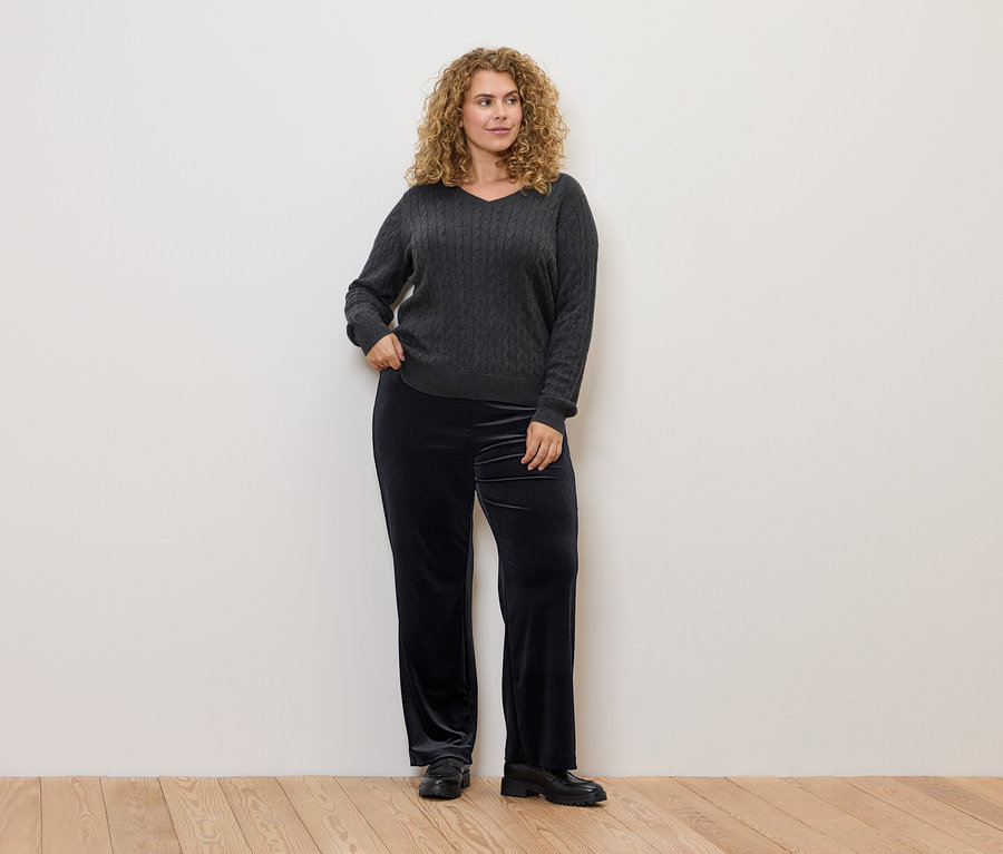 Frau mit lockigem Haar posiert in einem grauen Zopfmusterpullover und einer schwarzen Samthose.