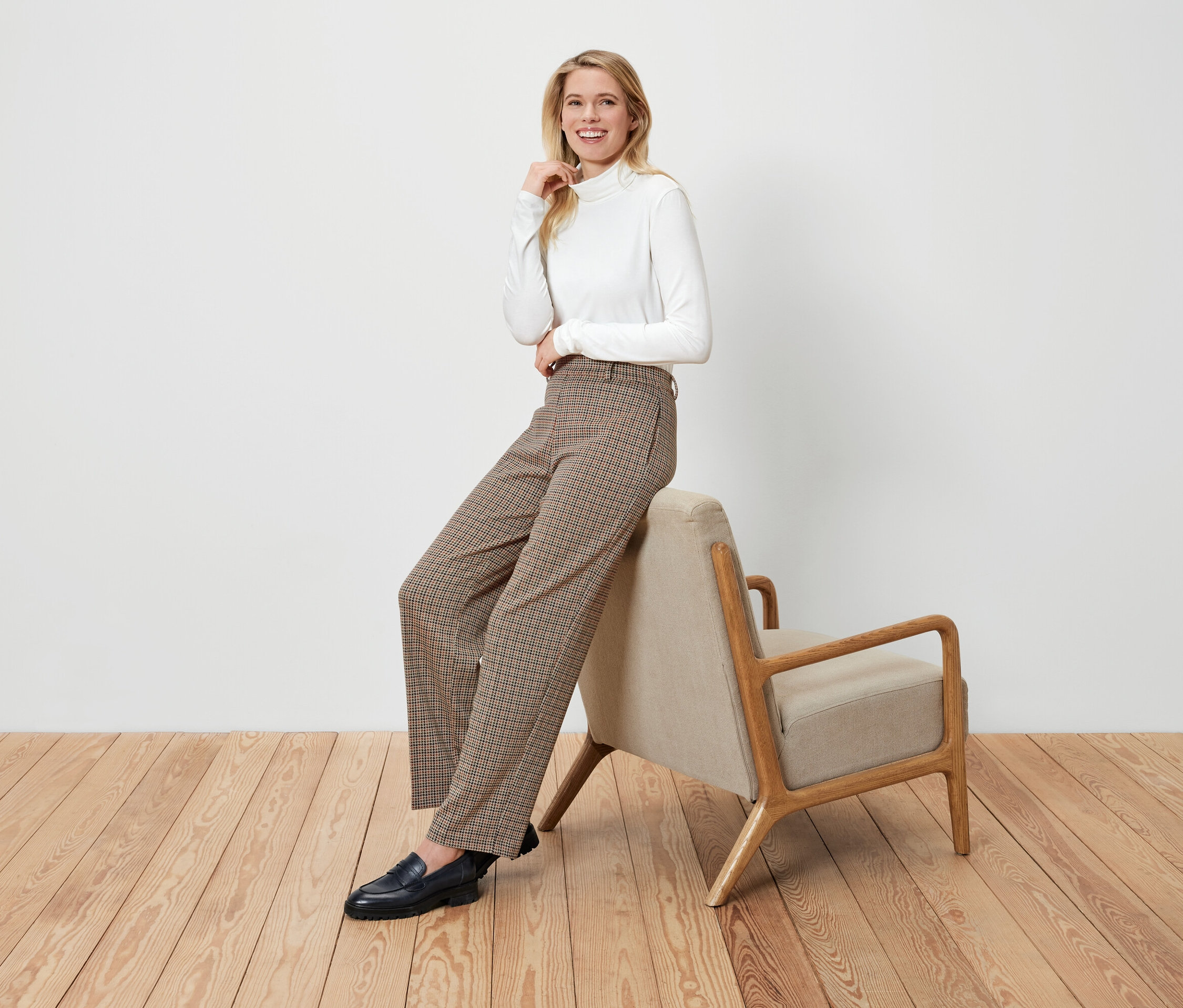 Blonde Frau sitzt auf einem beige Sessel, trägt einen weißen Rollkragenpullover, braune karierte Hosen und schwarze Slipper.