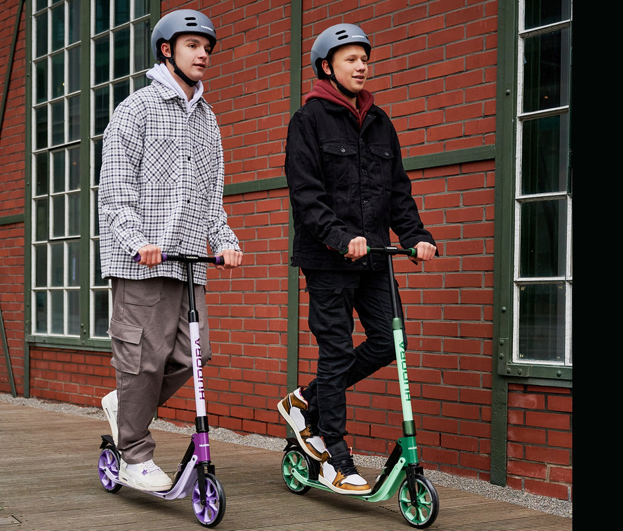 Zwei Teenager mit Helmen fahren auf HUDORA BigWheel® 205 Rollern vor einem Backsteingebäude.