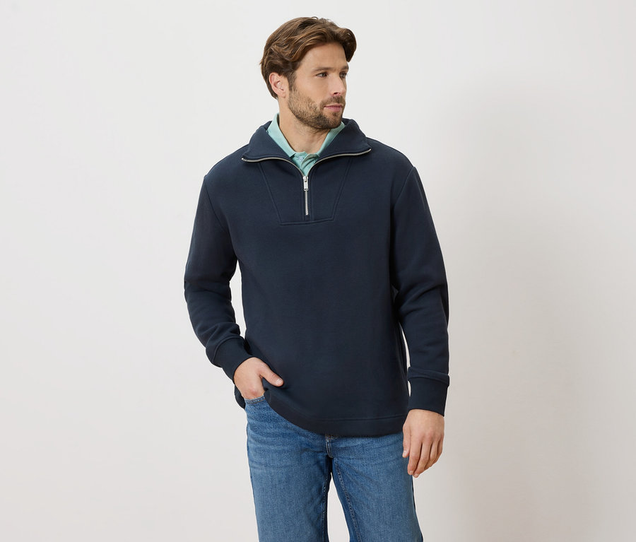 Mann mit marineblauem Quarter-Zip-Pullover und Jeans, Hand in der Tasche, Blick nach rechts.