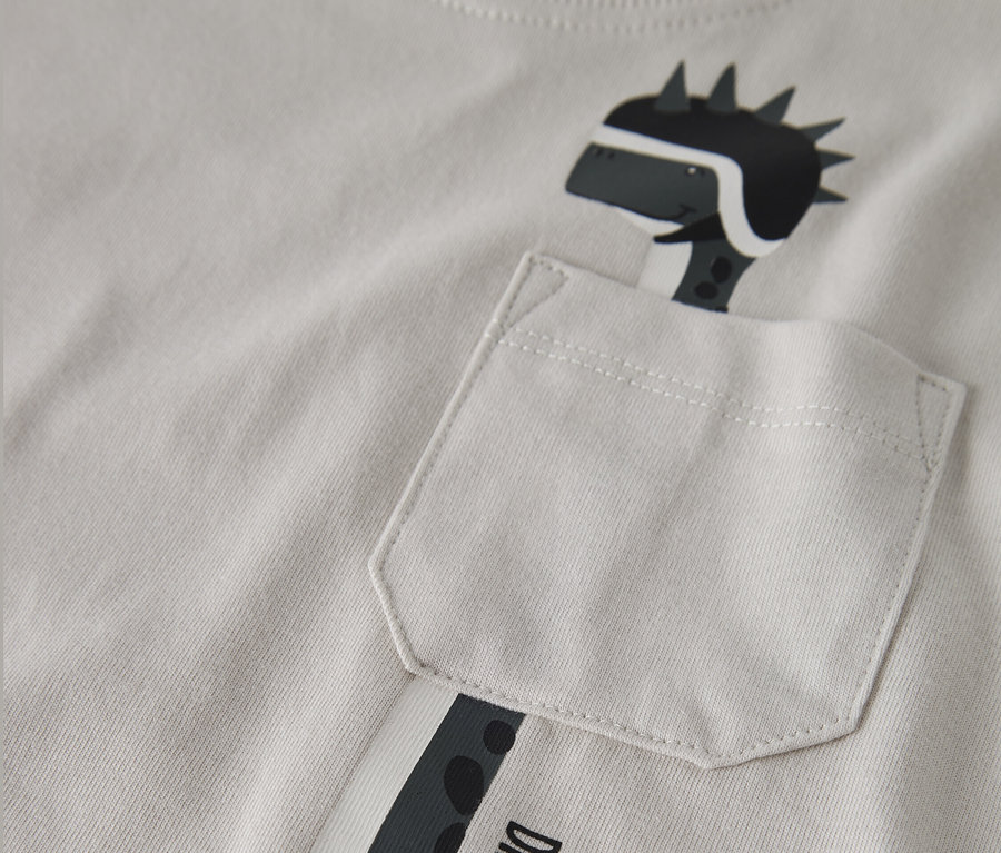 Detailansicht eines hellgrauen T-Shirts mit einer aufgedruckten Dinosaurierfigur, die einen Helm trägt und aus einer Brusttasche schaut. Auf dem T-Shirt steht der Text "Dino Racer Born to Race".