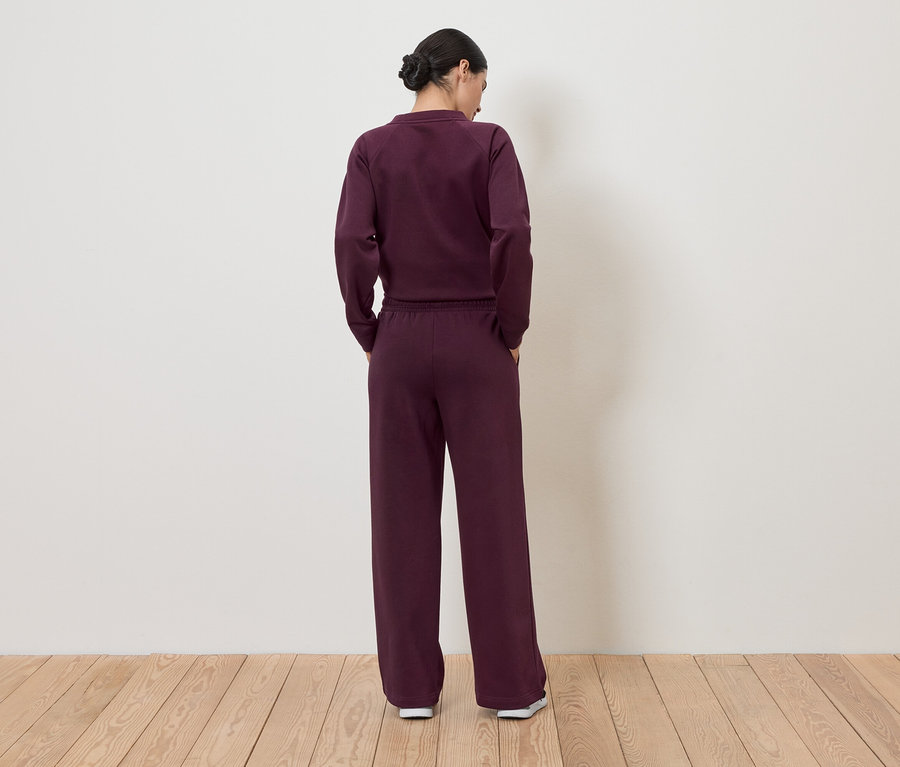 Rückansicht einer Frau mit hochgesteckten Haaren, die ein weinrotes Trainingsoutfit bestehend aus einem Oberteil und einer Hose trägt.