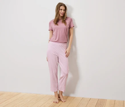 Eine Frau steht vor einer weißen Wand, sie trägt ein rosa T-Shirt und eine rosafarbene Hose mit Muster.