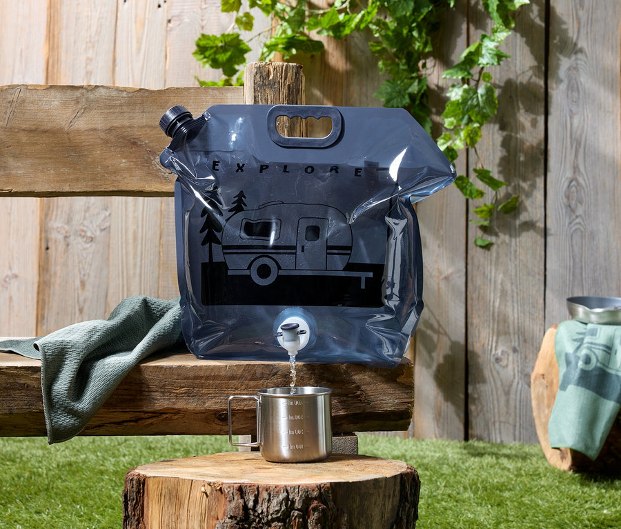 Wasser wird aus einem grauen Wasserbeutel mit Wohnwagen-Motiv in eine Tasse auf einem Baumstumpf gegossen.