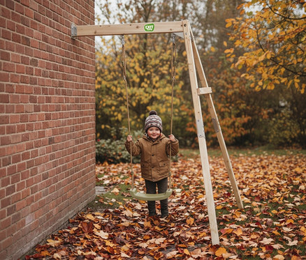 Ein lächelnder Junge mit Mütze schwingt auf einer Holzschaukel vor einer Backsteinmauer, umgeben von Herbstlaub.
