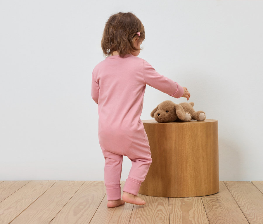Ein Kind in einem rosa Overall steht neben einem Holzhocker und berührt einen Stoffhund.