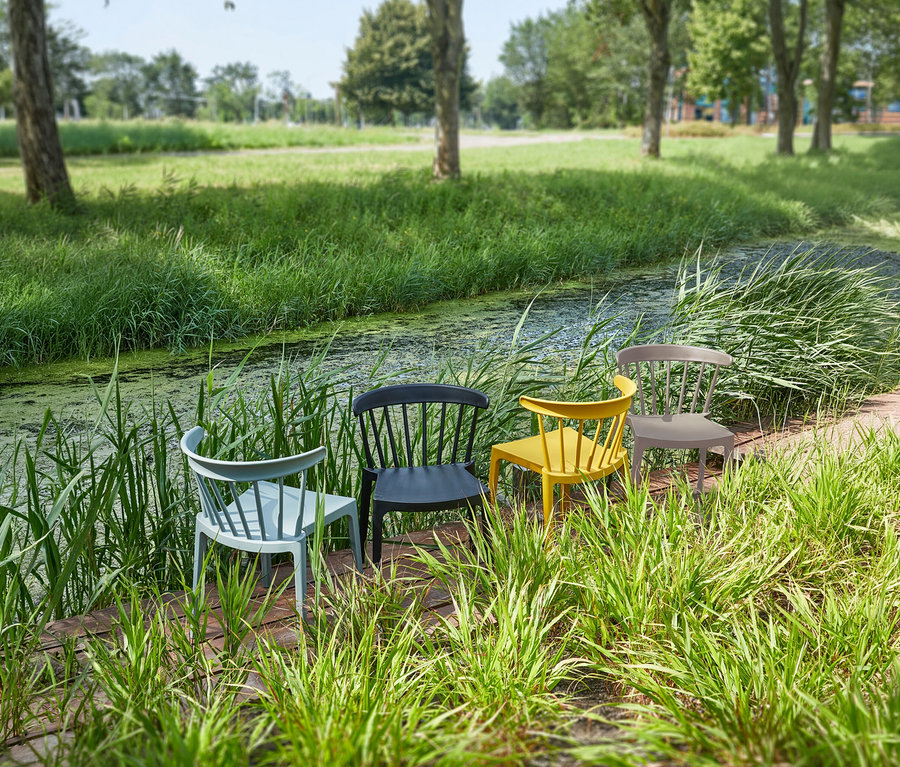 Vier WOOOD Gartenstühle in Blau, Schwarz, Karamell und Grau stehen auf einem Holzsteg vor einem Graben mit Gras im Hintergrund.