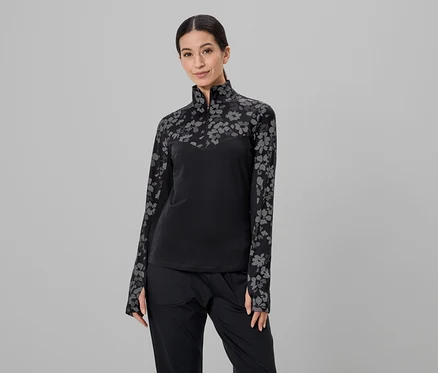 Frau mit Pferdeschwanz trägt schwarzes Oberteil mit Blumenmuster und schwarze Hose vor grauem Hintergrund.