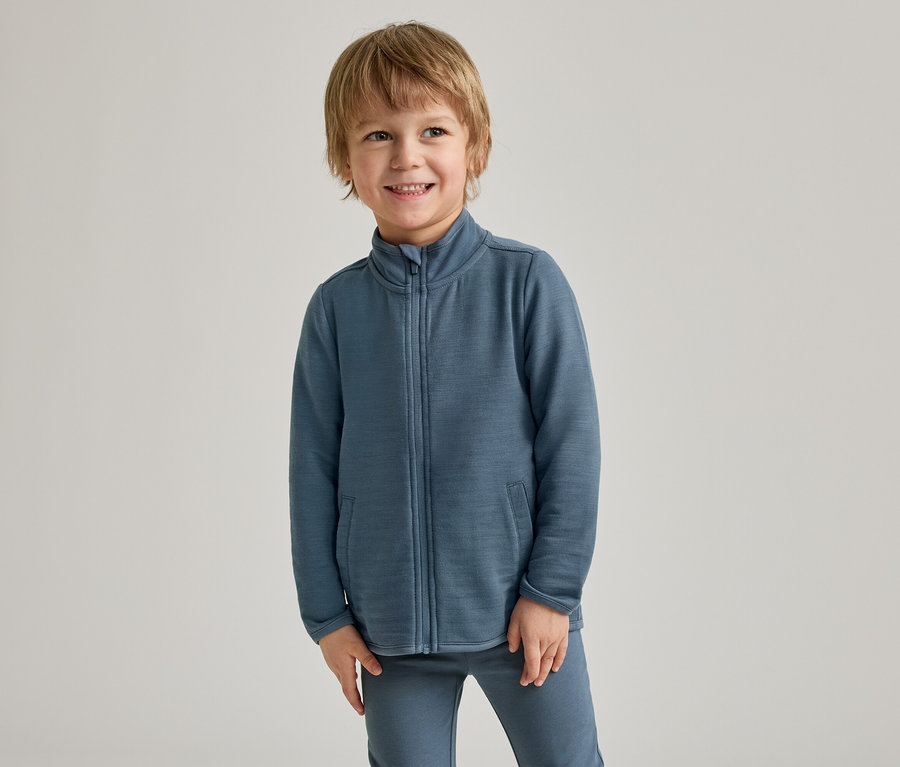 Ein kleiner Junge mit blondem Haar lächelt in einer blauen Jacke mit Reißverschluss und passender Hose vor hellem Hintergrund.