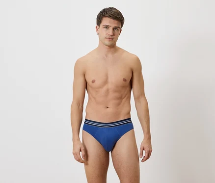 Mann mit freiem Oberkörper und blauen Slips mit blau-weißem Bund vor weißem Hintergrund.