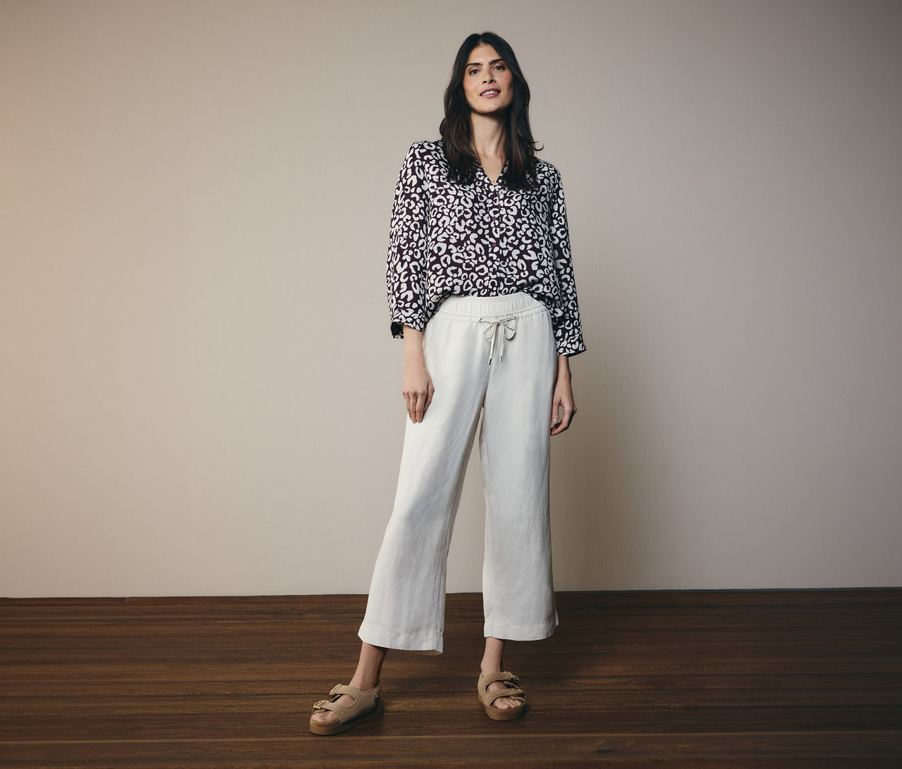 Frau trägt eine bordeauxrote CECIL Bluse mit hellem abstraktem Muster, hellbeige weite Cropped-Hose und beigefarbene Sandalen vor einer hellgrauen Wand.