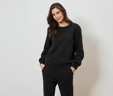 Frau trägt ein schwarzes Sweatshirt und eine Hose und posiert vor einem weißen Hintergrund.