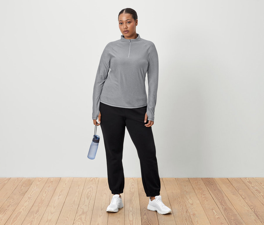 Eine Frau steht vor einer weißen Wand und hält eine blaue Wasserflasche in der Hand. Sie trägt ein graues Langarmshirt, schwarze Jogginghose und weiße Turnschuhe.