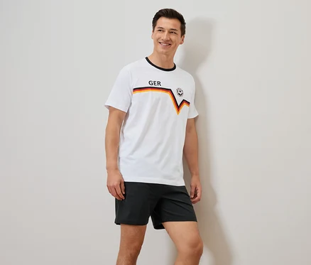 Mann mit einem weißen T-Shirt mit Deutschland-Flagge und schwarzen Shorts, der an einer Wand lehnt.