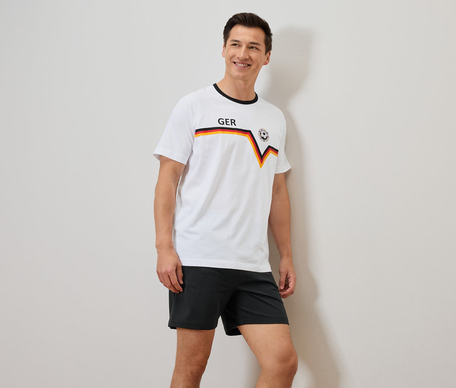 Mann mit einem weißen T-Shirt mit Deutschland-Flagge und schwarzen Shorts, der an einer Wand lehnt.