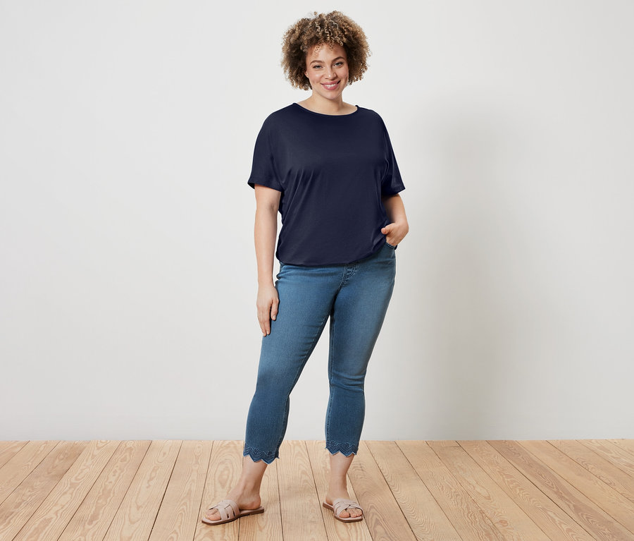 Frau mit lockigem Haar trägt ein marineblaues T-Shirt, blaue Jeans mit gewelltem Saum und Sandalen.