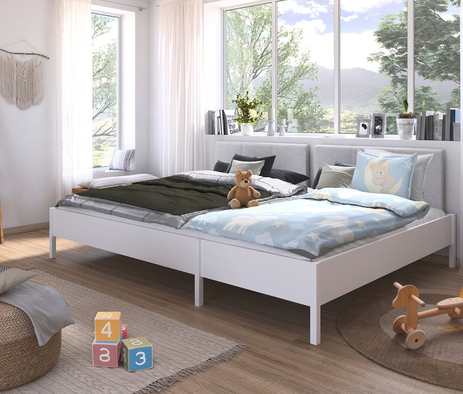 Zwei weiße Betten stehen in einem hellen Raum unter Fenstern. Auf einem Bett liegt ein Teddybär, das andere ist mit einer grünen Decke bedeckt.