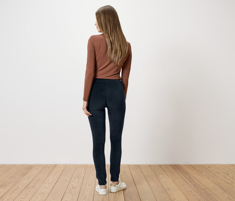 Frau mit braunem Oberteil und blauen Leggings, von hinten gesehen, vor weißer Wand und Holzboden.