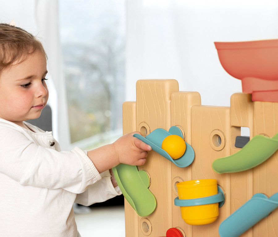Mädchen spielt mit der Smoby Life Activity Wall, indem es eine gelbe Kugel in eine blaue Schaufel legt.