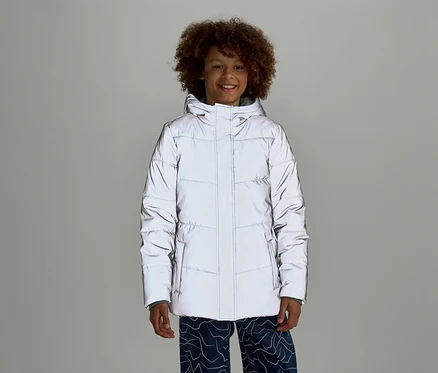 Ein lächelnder Junge mit lockigem Haar trägt eine reflektierende Pufferjacke mit Kapuze und blaue Hosen mit Muster vor einem grauen Hintergrund.