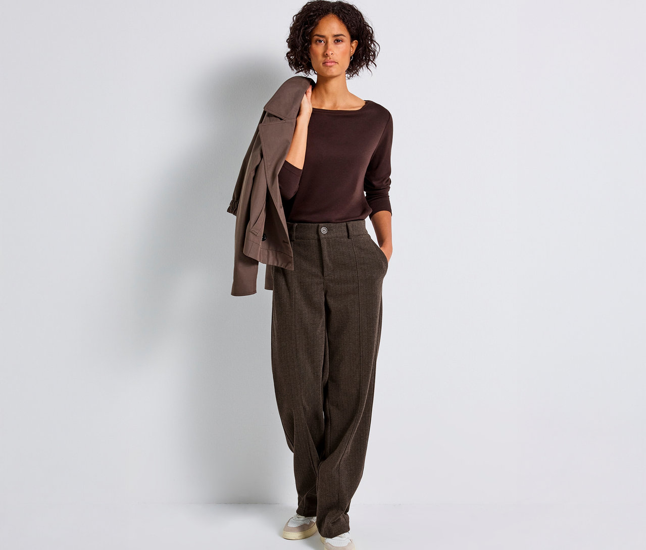 Frau steht und trägt eine braune Street One Hose Straight Leg, ein braunes Oberteil und eine Jacke über der Schulter.