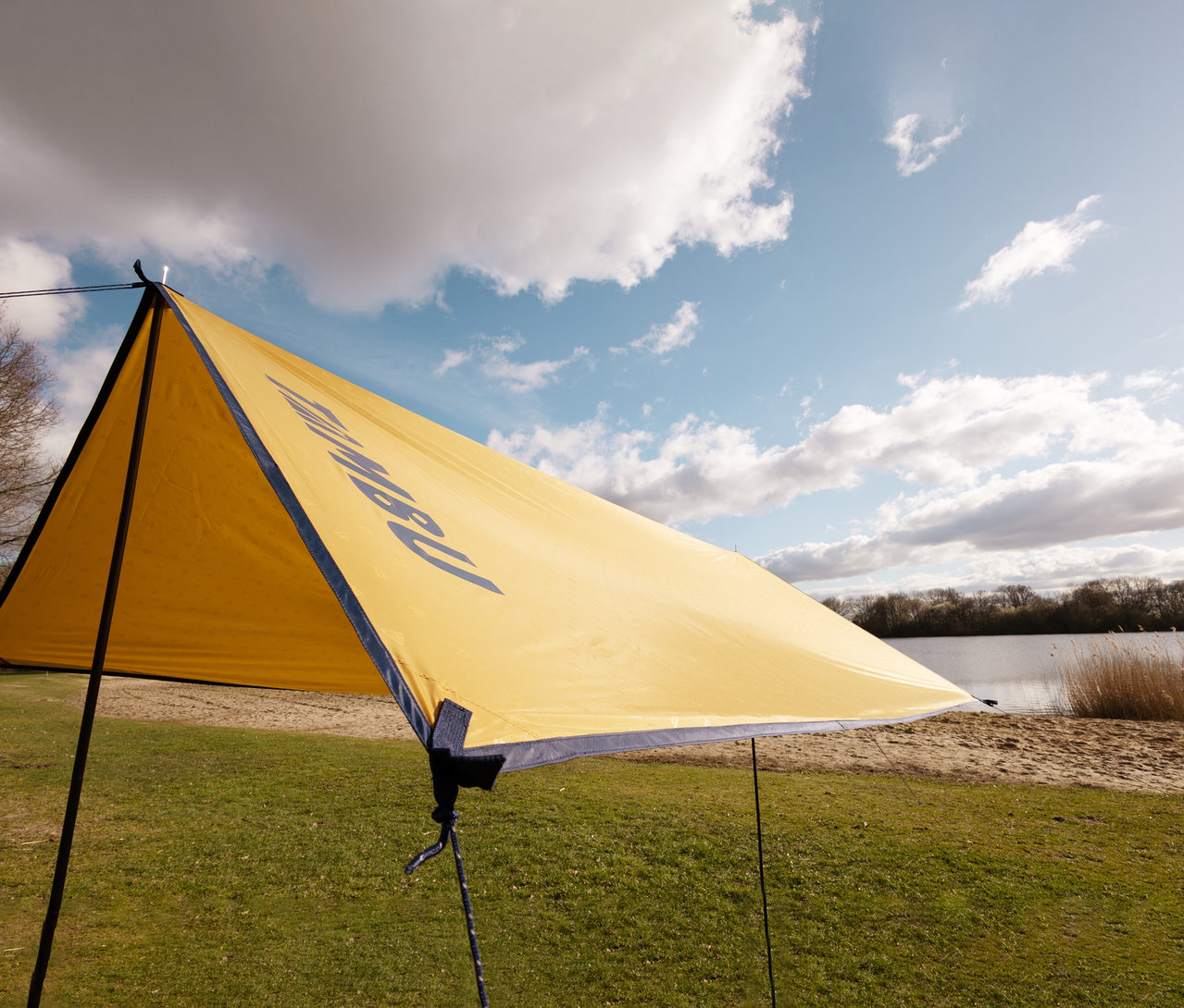 Auf einer Wiese steht ein gelbes Tambu »CHADA« Tarp/Sonnensegel, im Hintergrund ein See und bewölkter Himmel.