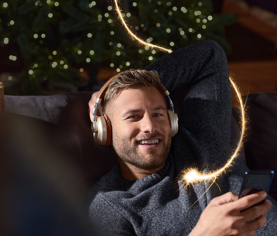 Lächelnder Mann liegt mit On-Ear-Bluetooth®-Kopfhörer auf Sofa, hält Handy in der Hand. Im Hintergrund ein Weihnachtsbaum.