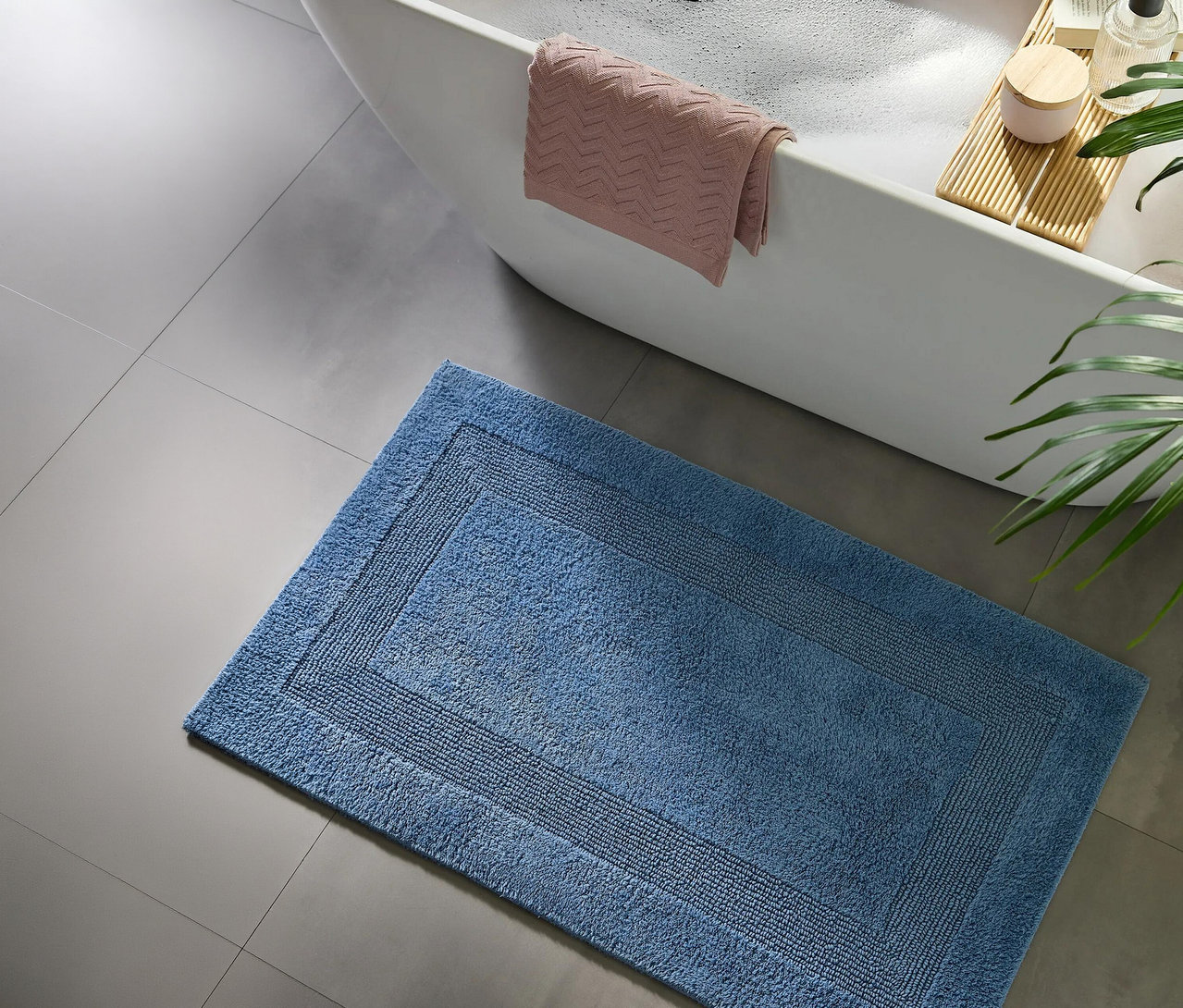 Blauer Badteppich auf grauen Fliesen neben einer weißen Badewanne mit rosa Handtuch und Badebrett.
