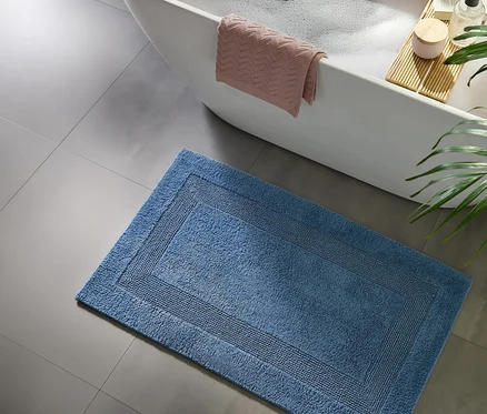 Blauer Badteppich auf grauen Fliesen neben einer weißen Badewanne mit rosa Handtuch und Badebrett.