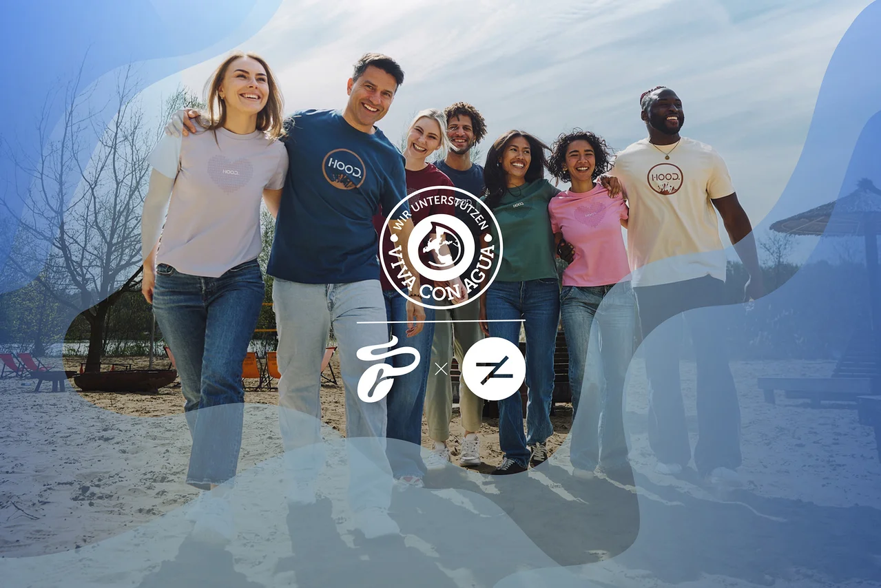 Sechs lächelnde Personen, drei Männer und drei Frauen, stehen Arm in Arm an einem Sandstrand. Sie tragen verschiedenfarbige HOOD.-T-Shirts und Jeans und blicken nach rechts. Ein „Wir unterstützen Viva con Agua“-Logo ist zentriert.