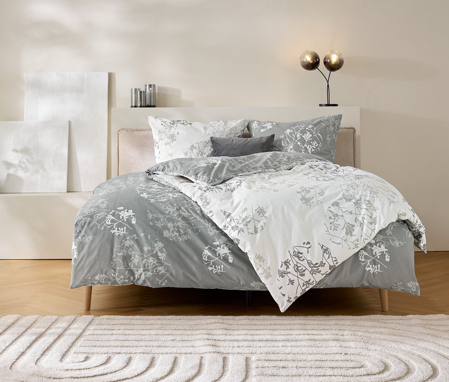 Ein Bett mit grauer Renforcé-Bettwäsche mit floralem Muster in Grau und Weiß. Die Bettwäsche ist teilweise zurückgeschlagen und zeigt beide Seiten. Auf dem Bett liegen zwei Kissen und ein kleines Zierkissen. Links vom Bett stehen eine schwarze Vase mit Trockenblumen und zwei weiße Kunstobjekte. Rechts steht eine Lampe mit zwei Glaskugeln. Vor dem Bett liegt ein heller Teppich mit einem kreisförmigen Muster.