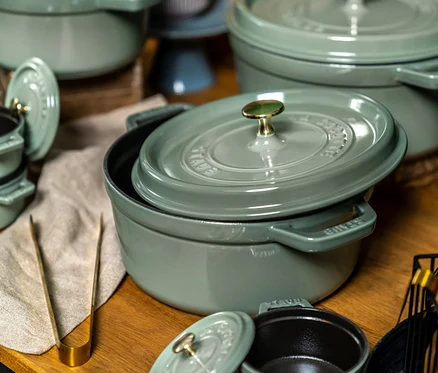 Eine Ansammlung grüner gusseiserner Töpfe und Auflaufformen auf einer Holzoberfläche. Im Vordergrund steht ein großer Topf, dessen Deckel leicht schräg aufliegt und das dunkle Innere zeigt. Goldene Zangen liegen links auf einem beigen Tuch.