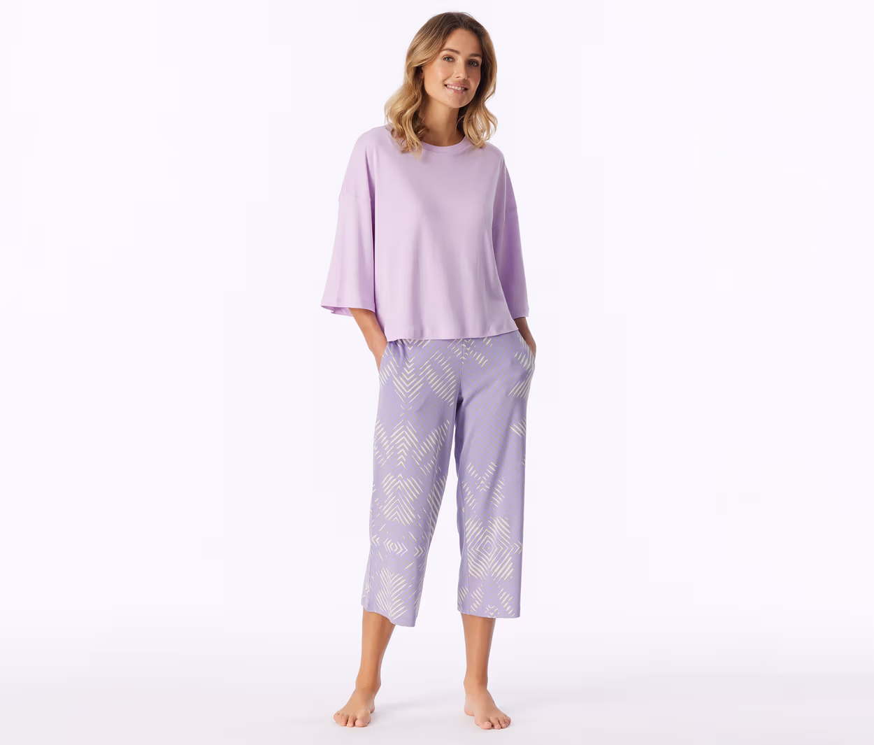 Lachende Frau steht barfuß, trägt ein hellviolettes Kurzarmshirt und hellviolette Caprihose mit weißem geometrischem Muster, Hände in den Taschen.