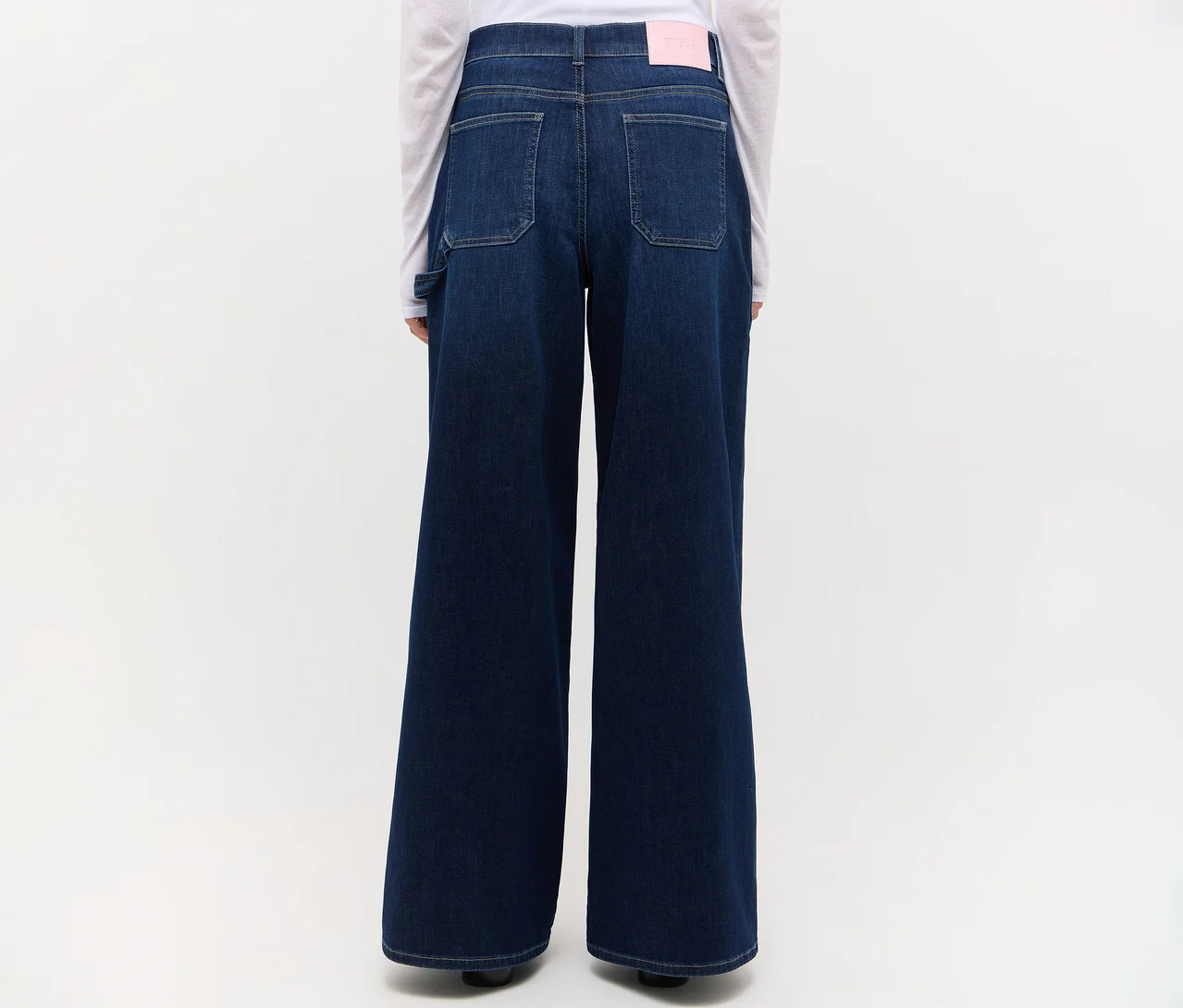 Rückenansicht einer Person in dunkelblauen, weit geschnittenen Jeans mit Gesäßtaschen und einem rosa Etikett am Bund, kombiniert mit einem weißen Langarmshirt.