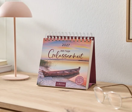 Ein 2027 Schreibtischkalender mit "365 Tage Gelassenheit" und einem Boot am Seeufer bei Sonnenuntergang steht auf einem Holztisch. Daneben eine rosa Lampe, Bücher und eine Brille.
