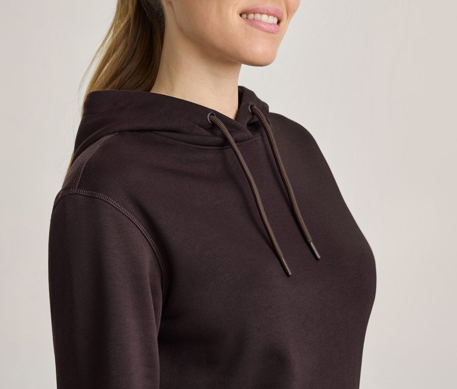 Frau in dunkelbraunem Kapuzenpullover mit Kordelzug, hellbraunes Haar im Pferdeschwanz, lächelt.