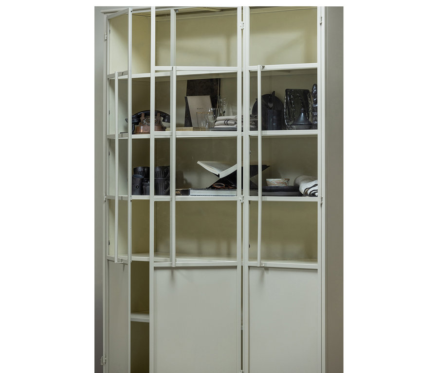Ein hoher, hellbeiger Vitrinenschrank mit Glastüren und massiven Bodentüren, der verschiedene Gegenstände präsentiert. Die Regale enthalten ein Vintage-Telefon, Gläser, gefaltete Textilien, dunkle Tassen, ein offenes Buch auf einem Ständer und Dekorationsgegenstände.